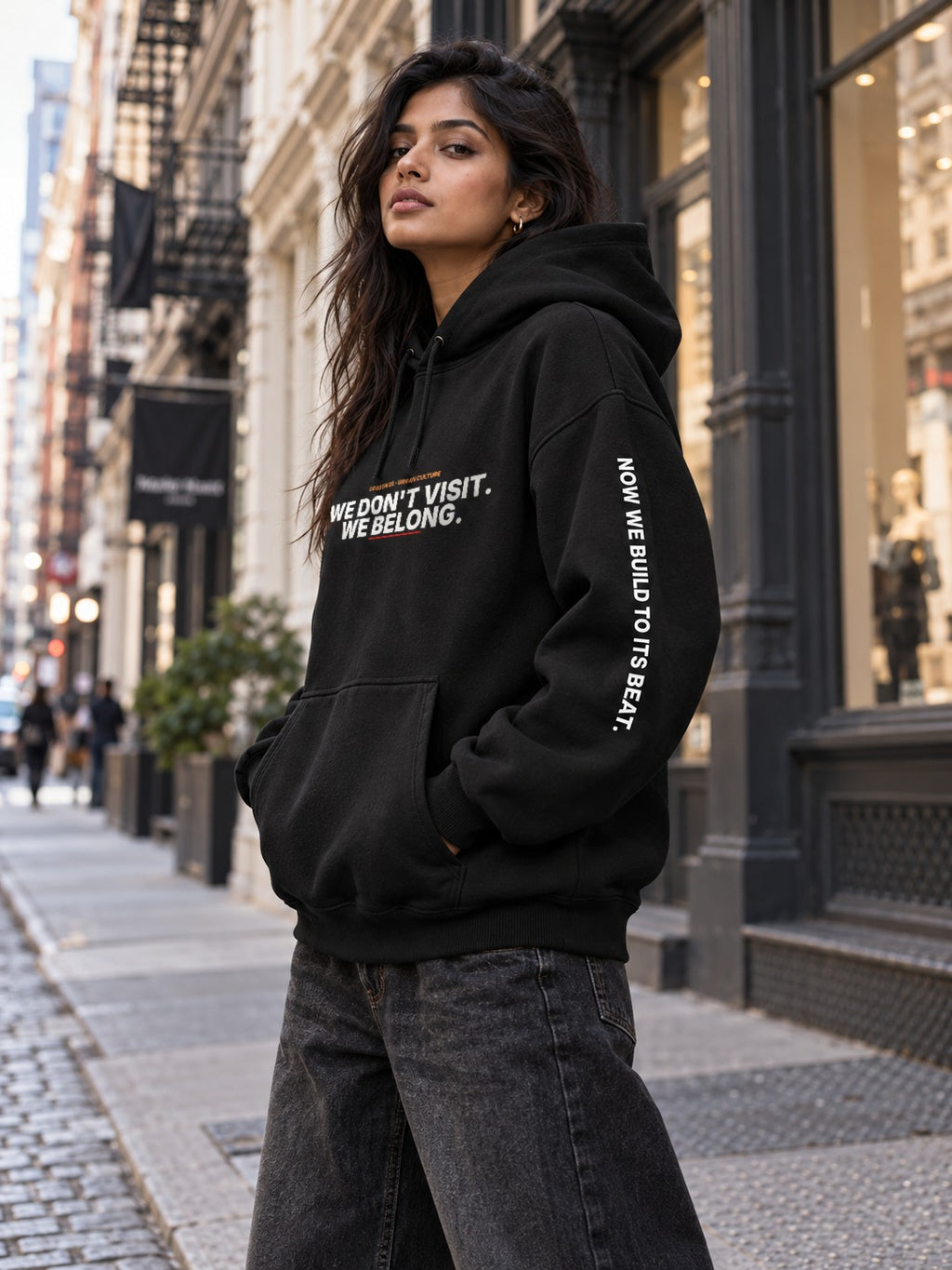 Woman with long dark hair on a SoHo sidewalk, turned slightly to the side in a black hoodie that reads ‘We Don’t Visit. We Belong.’ with ‘Now We Build to Its Beat.’ down the right sleeve, one hand in the front pocket