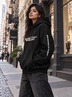 Woman with long dark hair on a SoHo sidewalk, turned slightly to the side in a black hoodie that reads ‘We Don’t Visit. We Belong.’ with ‘Now We Build to Its Beat.’ down the right sleeve, one hand in the front pocket