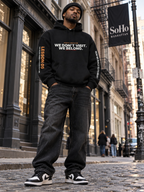 Low-angle shot of a man in a black beanie standing on a SoHo cobblestone street under a SoHo Manhattan banner, wearing a black hoodie that reads ‘We Don’t Visit. We Belong.’ with ‘Lesson 05’ on the left sleeve and ‘Now We Build to Its Beat.’ on the right, black jeans, and black-and-white sneakers