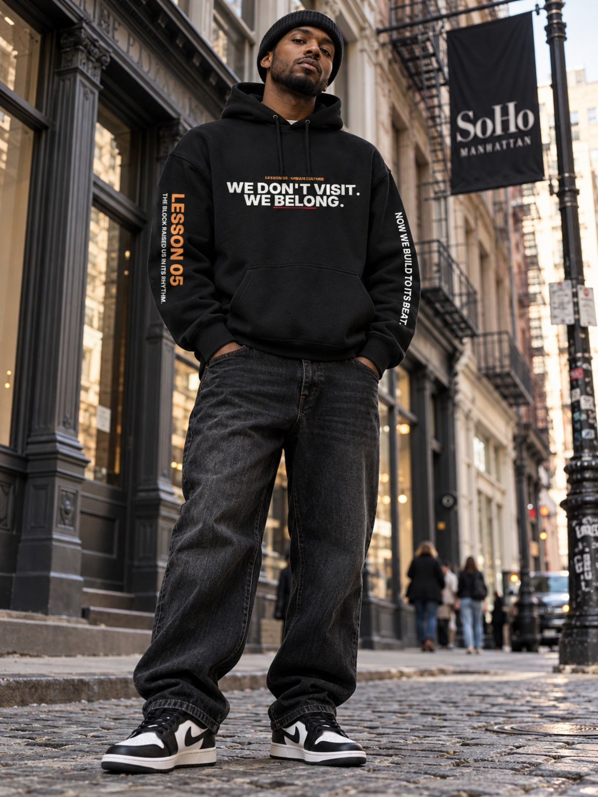 Low-angle shot of a man in a black beanie standing on a SoHo cobblestone street under a SoHo Manhattan banner, wearing a black hoodie that reads ‘We Don’t Visit. We Belong.’ with ‘Lesson 05’ on the left sleeve and ‘Now We Build to Its Beat.’ on the right, black jeans, and black-and-white sneakers