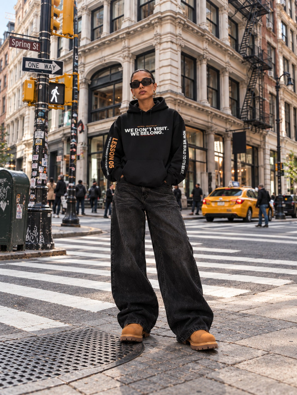 Woman in sunglasses standing at the Spring Street crosswalk with a yellow taxi behind her, wearing a black hoodie that reads ‘We Don’t Visit. We Belong.’ with ‘Lesson 05’ on the left sleeve and ‘Now We Build to Its Beat.’ on the right, wide-leg black jeans, and tan boots.
