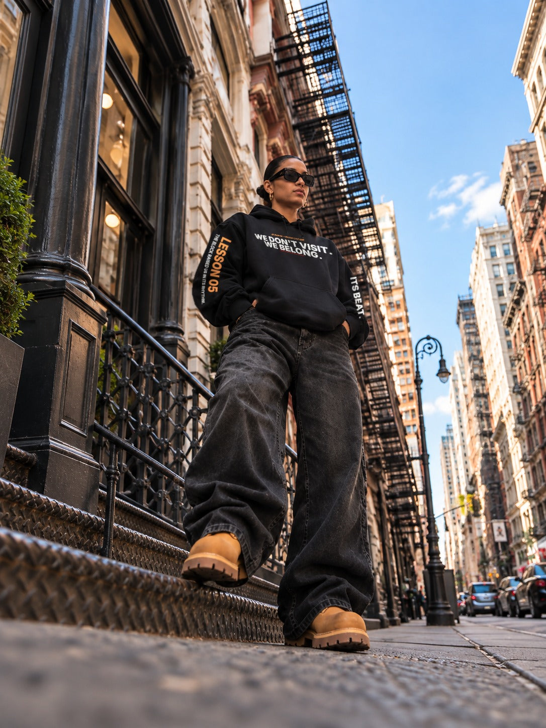 Low-angle shot of a woman in sunglasses stepping down a stoop between fire escapes under a blue sky, wearing a black hoodie that reads ‘We Don’t Visit. We Belong.’ with ‘Lesson 05’ on the left sleeve, wide-leg black jeans, and tan boots