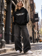 Woman with long blonde hair on a SoHo cobblestone street, wearing a black hoodie that reads ‘We Don’t Visit. We Belong.’ with ‘Lesson 05’ on the left sleeve and ‘Now We Build to Its Beat.’ on the right, wide-leg black jeans, and white sneakers