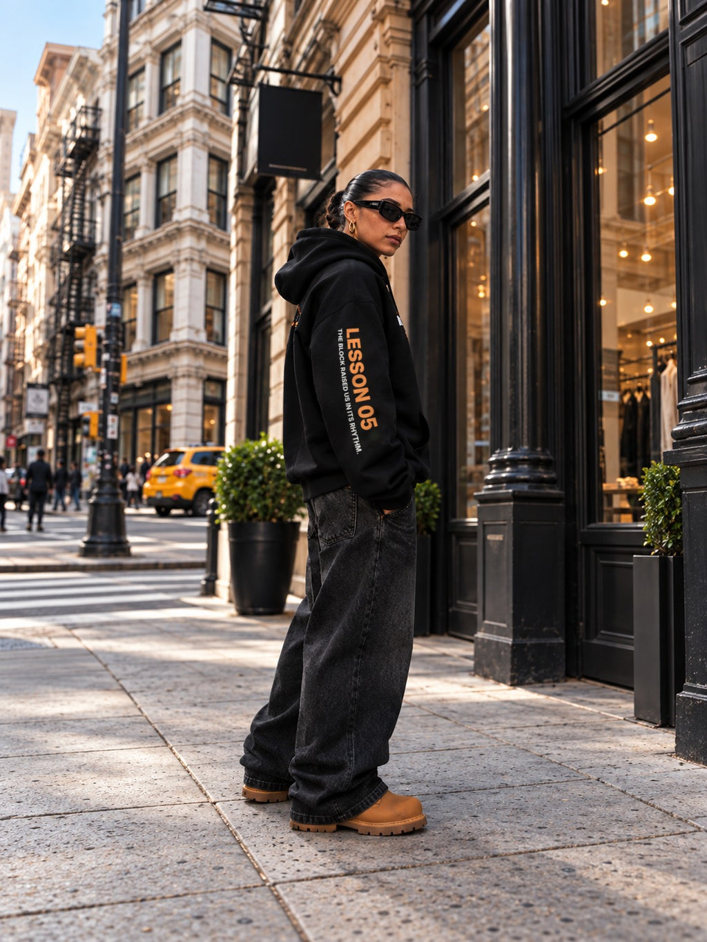 Side view of a woman in sunglasses on a SoHo sidewalk with a yellow taxi behind her, showing the left sleeve of a black hoodie printed with ‘Lesson 05’ and ‘The Block Raised Us in Its Rhythm.’, with wide-leg black jeans and tan boots.