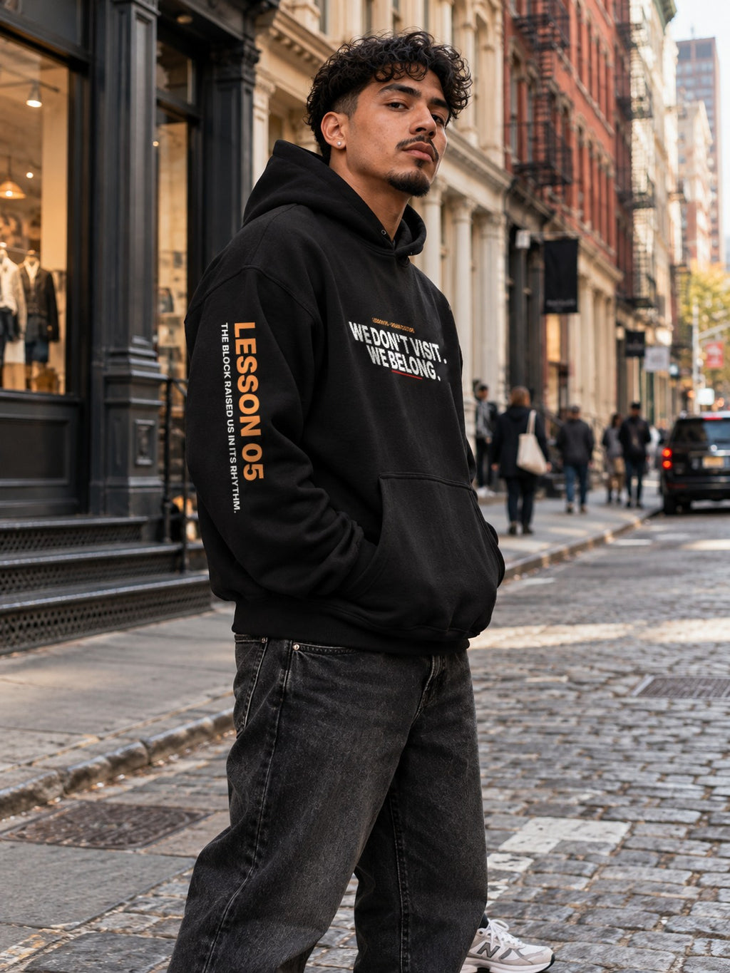 Man with curly hair on a SoHo cobblestone street, angled to the side with his hands in the pocket of a black hoodie that reads ‘We Don’t Visit. We Belong.’ with ‘Lesson 05’ and ‘The Block Raised Us in Its Rhythm.’ down the left sleeve.