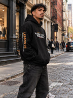 Man with curly hair on a SoHo cobblestone street, angled to the side with his hands in the pocket of a black hoodie that reads ‘We Don’t Visit. We Belong.’ with ‘Lesson 05’ and ‘The Block Raised Us in Its Rhythm.’ down the left sleeve.
