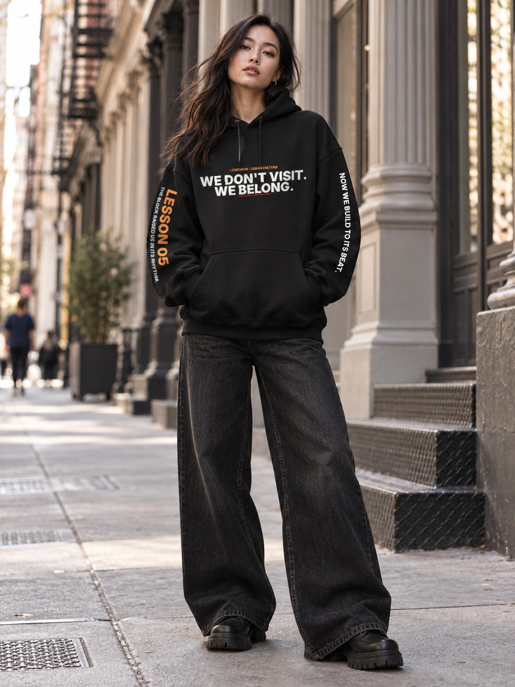 Woman with long dark hair standing on a sidewalk in front of stone columns, wearing a black hoodie that reads ‘We Don’t Visit. We Belong.’ with ‘Lesson 05’ on the left sleeve and ‘Now We Build to Its Beat.’ on the right, wide-leg black jeans, and black boots.