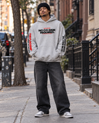 Male model wearing the gray BSchool Flagship Hoodie with black baggy jeans, sneakers, and a beanie on an NYC sidewalk. Front-facing streetwear shot highlights the “NO CO-SIGN REQUIRED.” chest graphic and printed sleeves.