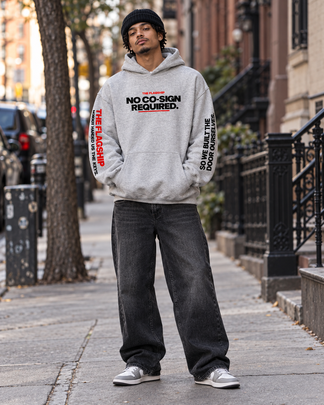 Male model wearing the gray BSchool Flagship Hoodie with black baggy jeans, sneakers, and a beanie on an NYC sidewalk. Front-facing streetwear shot highlights the “NO CO-SIGN REQUIRED.” chest graphic and printed sleeves.
