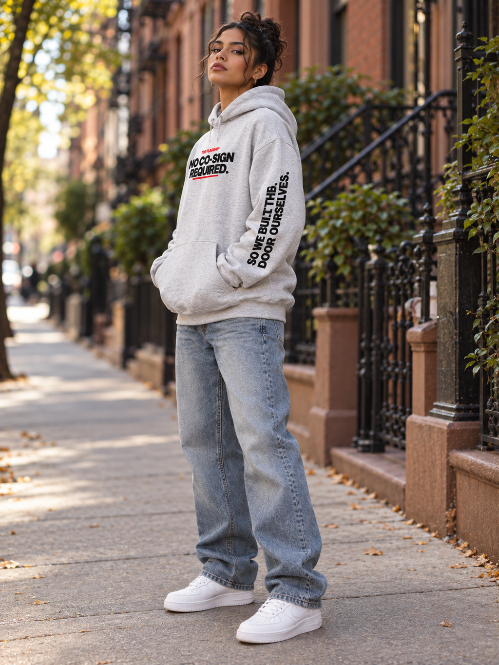 Latina woman modeling the gray BSchool Flagship Hoodie on an NYC brownstone sidewalk. Three-quarter side view highlights the black “SO WE BUILT THE DOOR OURSELVES.” sleeve graphic.