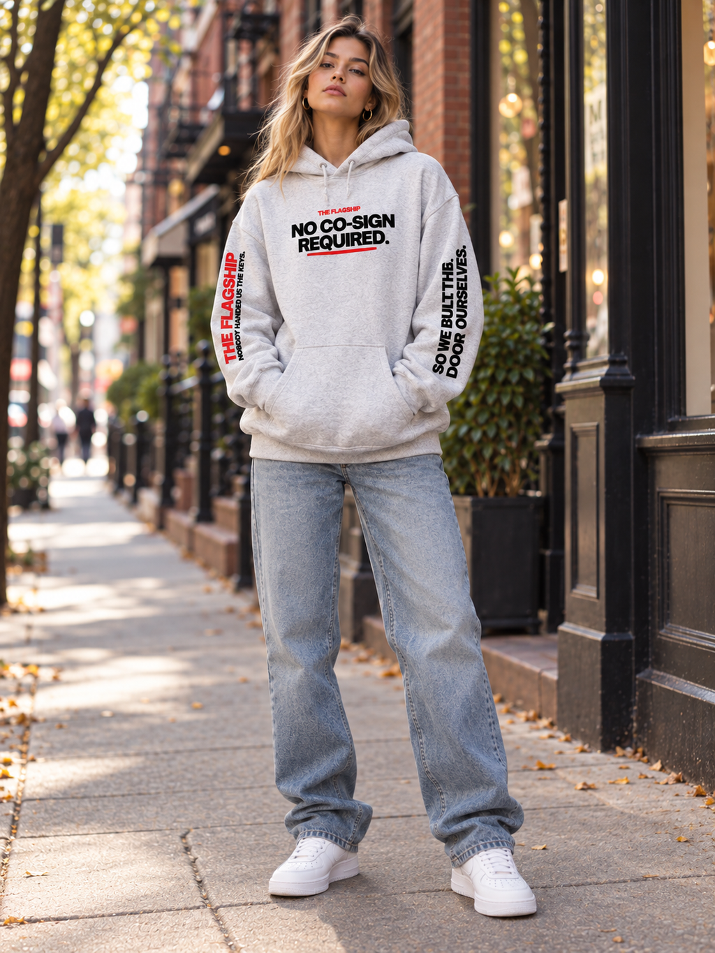 Blonde female model wearing the gray BSchool Flagship Hoodie with baggy jeans and white sneakers on an NYC sidewalk. Front-facing editorial shot showcases the “NO CO-SIGN REQUIRED.” chest graphic and both printed sleeves.
