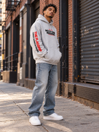 Black male model wearing the gray BSchool Flagship Hoodie on an industrial NYC street. Three-quarter angle highlights the red “THE FLAGSHIP” sleeve graphic, chest print, and relaxed streetwear fit.