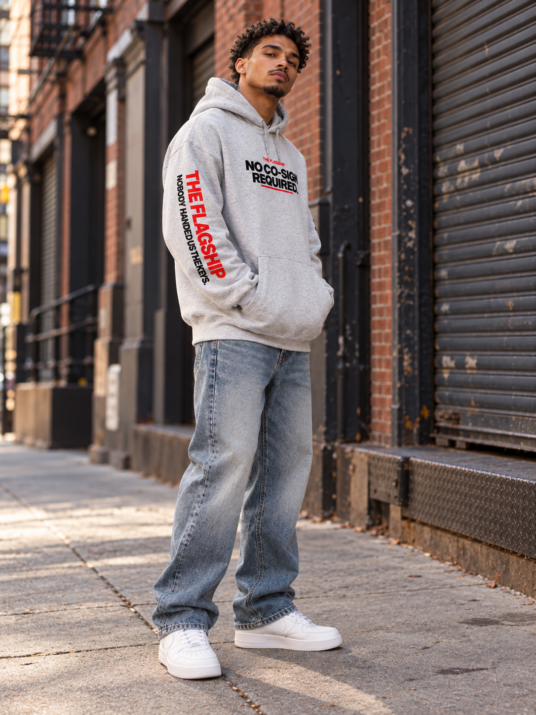 Black male model wearing the gray BSchool Flagship Hoodie on an industrial NYC street. Three-quarter angle highlights the red “THE FLAGSHIP” sleeve graphic, chest print, and relaxed streetwear fit.