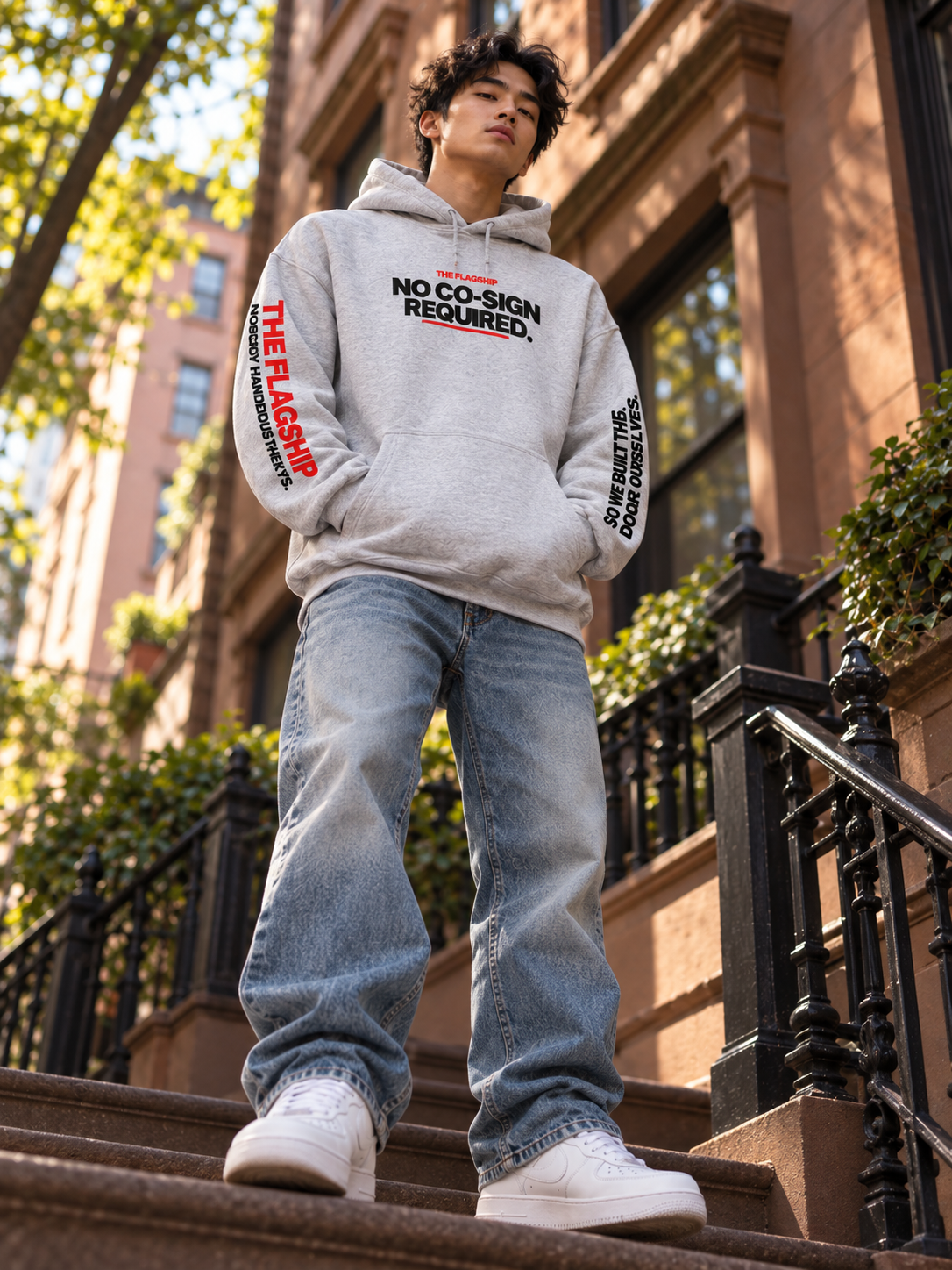Asian male model wearing the gray BSchool Flagship Hoodie on NYC brownstone steps. Low-angle streetwear photograph showcases the chest graphic and red and black sleeve prints.